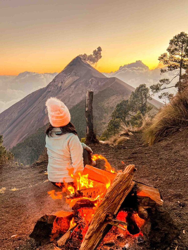 woman stands in front of campfire with Volcan Fuego in background with a sunset sky