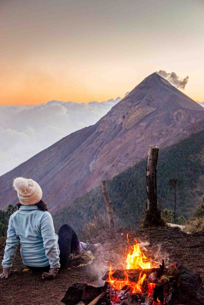 Woman sits in front of campfire against Volcan Fuego at sunset