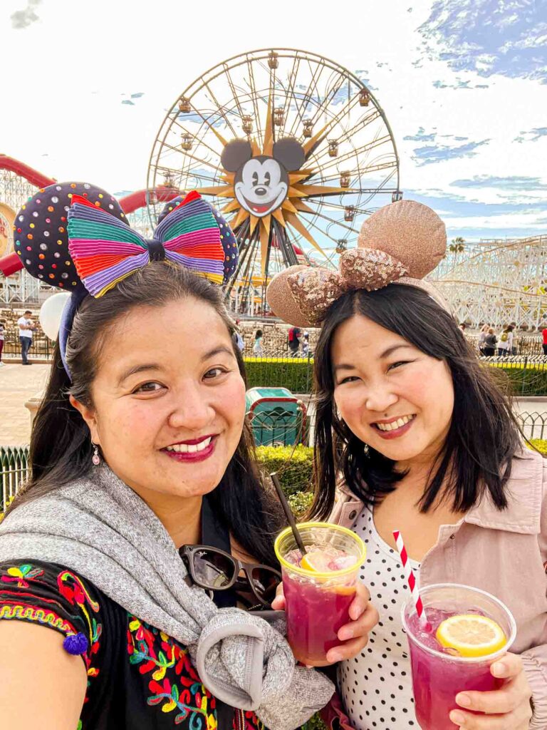 Selfie of two women wearing mouse ears and holding cocktails in front of Mickey ferris wheel